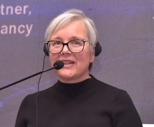 Caroline Bremner speaks into a headset microphone while presenting on stage, wearing a black top against a large screen backdrop.