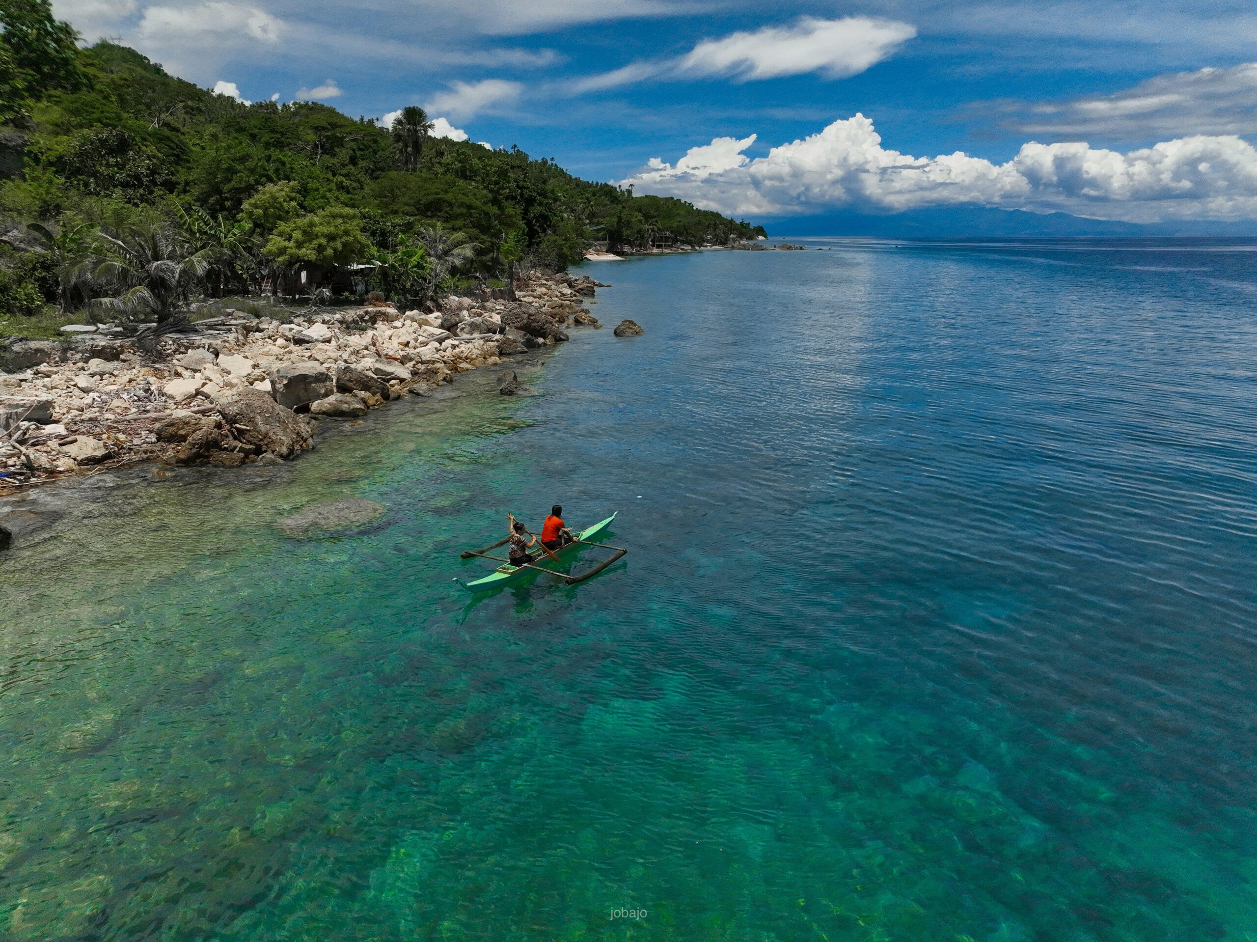 Two people paddling a kayak in clear turquoise water near a rocky coastline.