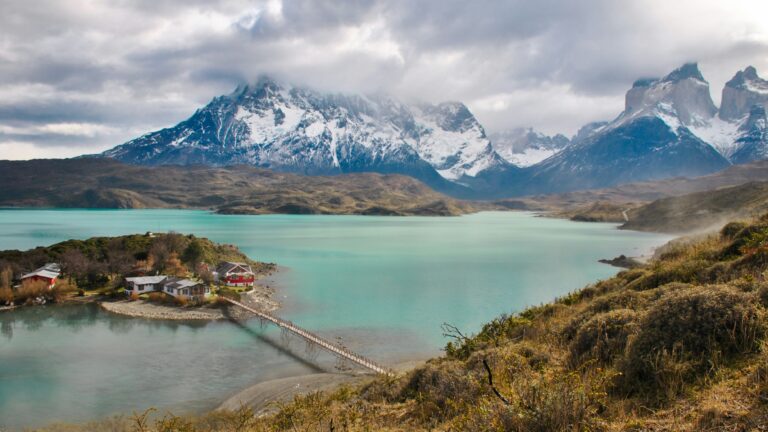 Turquoise lake with a small cluster of buildings connected by a narrow bridge, set against snow-capped mountains.