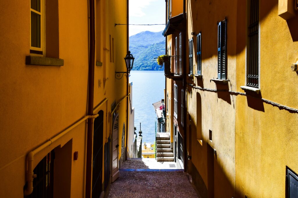 Narrow alleyway between yellow buildings leading down to a lake with mountains in the distance.