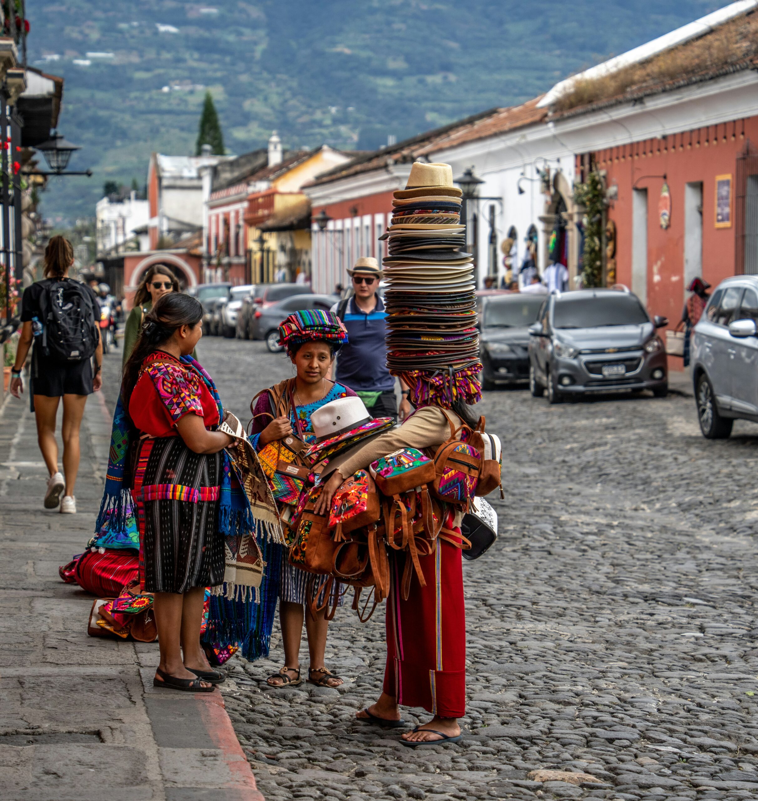 Street vendor in traditional clothing selling stacked hats to women on a cobbled street.