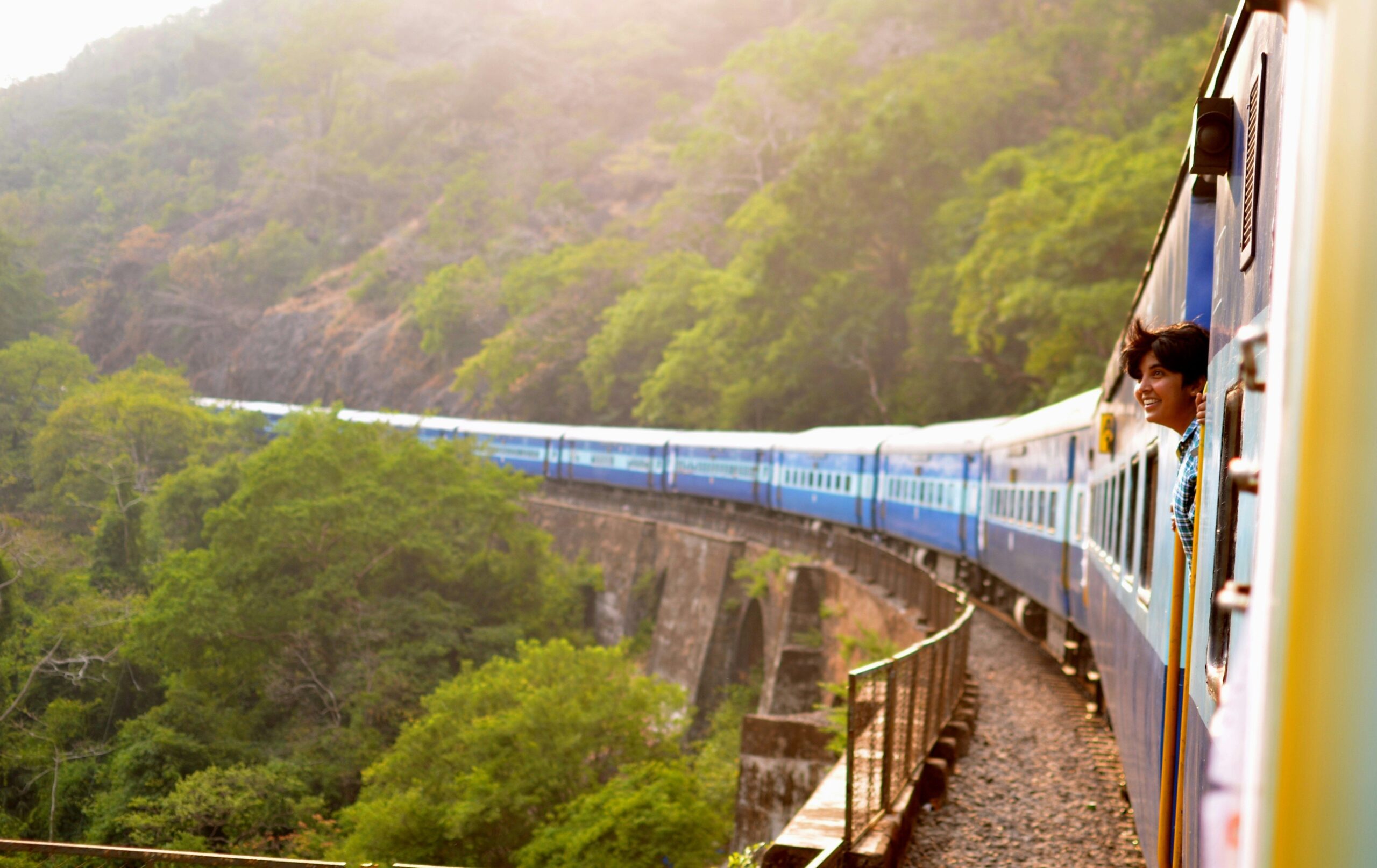 Passenger train crossing a curved bridge through forested hills in India, with a traveller leaning out of the carriage window.