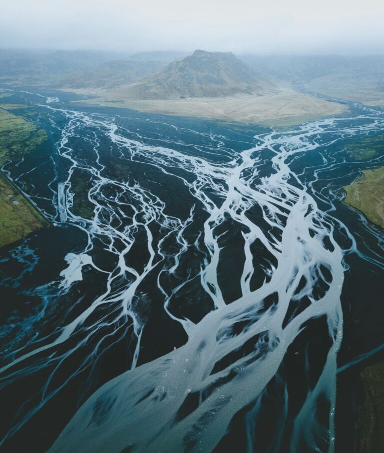 Aerial view of a braided river with pale blue channels winding across dark volcanic sediment towards distant mountains.